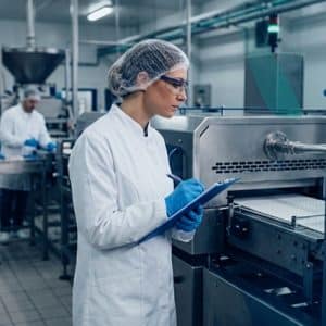 Una auditora de inocuidad alimentaria con bata blanca, cofia, gafas de seguridad y guantes azules toma notas en una tabla técnica mientras inspecciona una línea de producción con maquinaria de acero inoxidable.